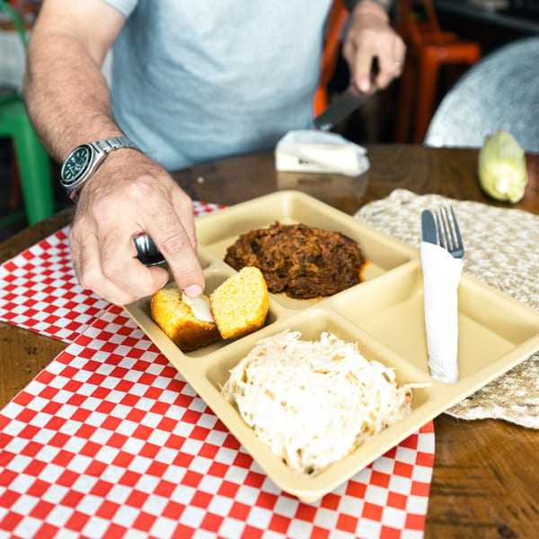 A person buttering cornbread on a Smoke Shack BBQ tray with pulled pork and coleslaw.