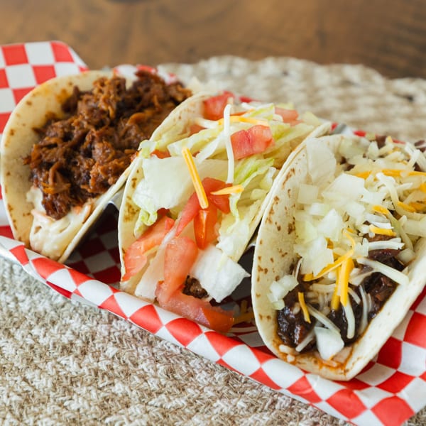 Three Smoke Shack BBQ tacos with various toppings, served in a red and white checkered tray.