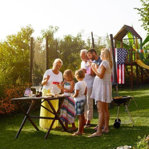A family gathers around a table and grill in a sunny backyard for a BBQ, with an American flag.