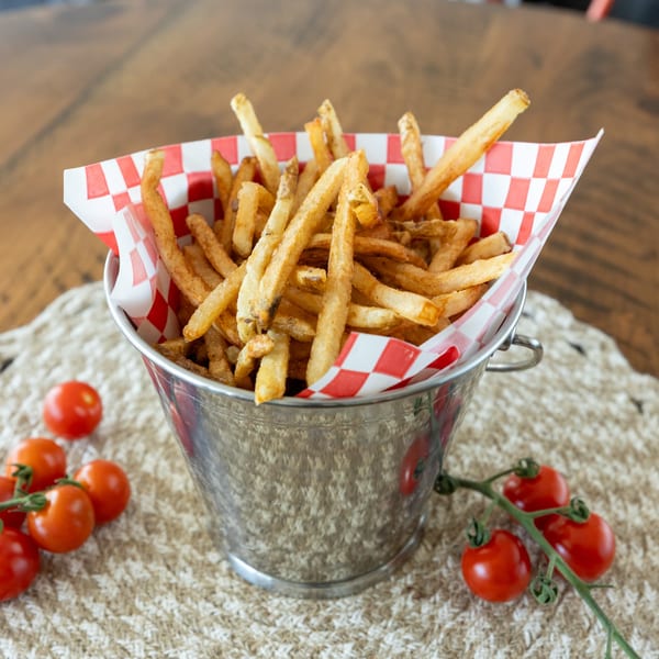 Crispy french fries in a metal bucket with red and white checkered paper, alongside cherry tomatoes.