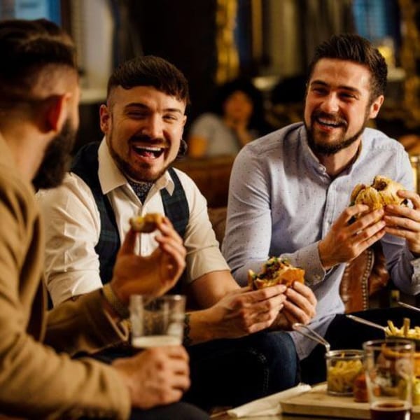 Three men laughing and eating Smoke Shack BBQ burgers and fries with beers.