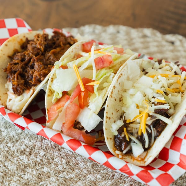 Three BBQ tacos in a red and white checkered holder, filled with meat, cheese, lettuce, and tomatoes.