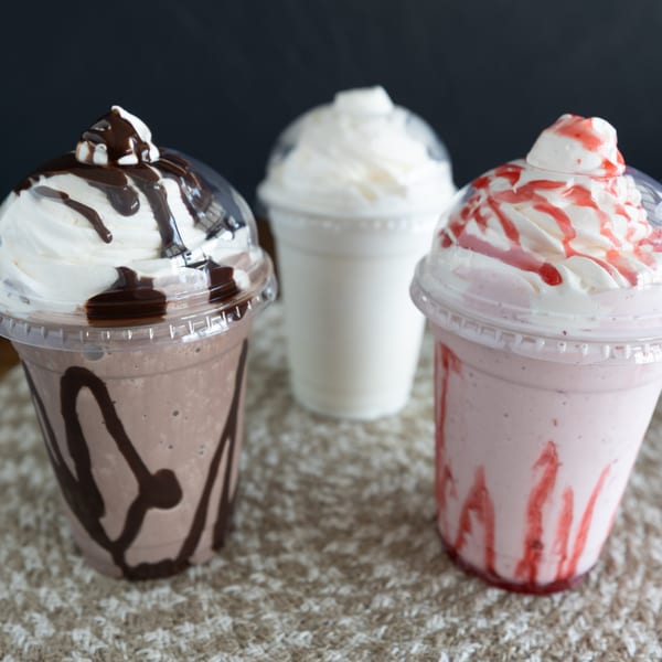 Three milkshakes: chocolate with fudge, vanilla, and strawberry with strawberry syrup, on a textured surface.