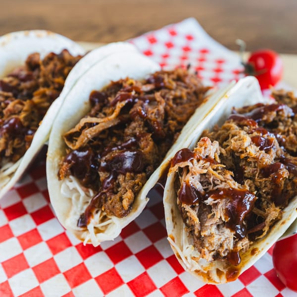 Three Smoke Shack BBQ pulled pork tacos with sauce and coleslaw on a red and white checkered paper.