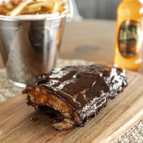 Smoke Shack BBQ ribs with dark sauce on a wooden board, with fries and a drink in the background.